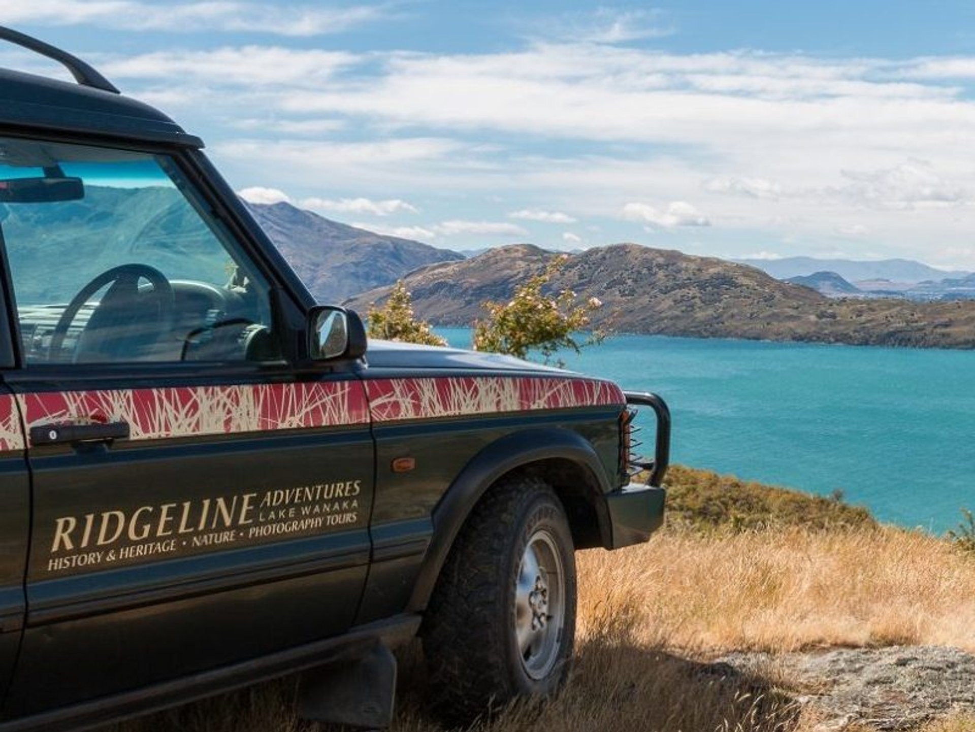 Ridgeline Adventures SUV kijkt uit over het turquoise Lake Wanaka met bergen op de achtergrond in Nieuw-Zeeland