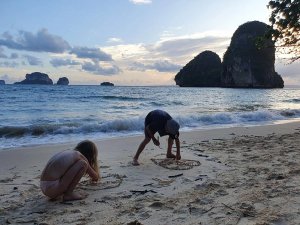 Jongen en meisje tekenen in het zand in Thailand. De zee golft achter hun en de lucht kleurt oranje van de zon en lichtblauw met lichte wolken boven de rotsformaties in zee.
