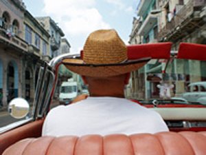 Persoon met strohoed rijdt in een rode cabrio door een stadsstraat gezien van achteren in Cuba
