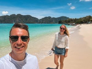 Twee reizigers wandelen lachend langs een tropisch zandstrand met turquoise zee en groene eilanden op de achtergrond. Blauwe lucht