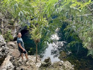 Een man staat op rotsachtige grond naast een helderblauw natuurlijk zwembad en de ingang van een grot, omringd door weelderige junglevegetatie in Cuba
