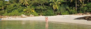 Volwassene staat met kind in ondiep zeewater bij een rustig strand met palmbomen en wit zand.