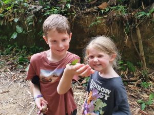 Twee kinderen bekijken een groene vlinder op de hand van een jongen in een bosrijke omgeving.