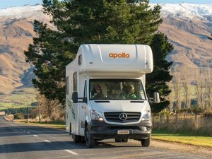Witte Apollo camper met twee personen rijdt over een weg met besneeuwde bergen en bomen op de achtergrond in een bergachtig natuurgebied