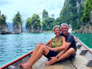 Twee reizigers zitten samen op de punt van een longtailboot op het water tussen kalksteenrotsen.