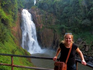 Vrouwelijke reiziger poseert langs een reling met op de achtergrond een hoge waterval, omringd door dichte junglebegroeiing aan beide zijden.
