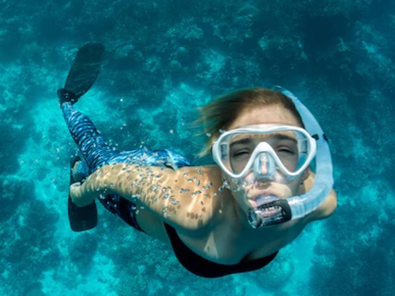 Vrouw snorkelt in helderblauw tropisch water, omringd door koraal en zonlicht dat door het wateroppervlak schijnt. Gezicht van de vrouw naar de camera.