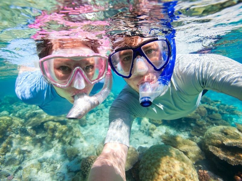 Twee snorkelende reizigers onder water boven kleurrijk koraalrif maken een selfie in helder tropisch water.