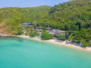 Azuurblauw water en een rustig strand, omringd door tropisch groen met een kleinschalig resort verscholen tussen de bomen.