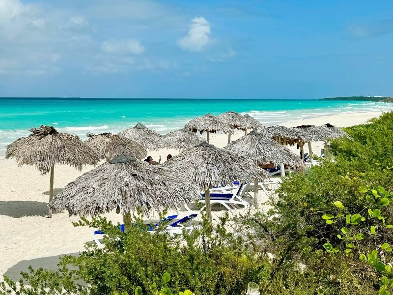Een tropisch strand met wit zand, turquoise water, rieten parasols, ligstoelen en groene struiken.