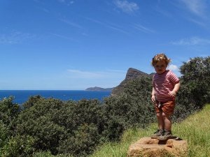 Peuter staat op een rots met uitzicht op een blauwe oceaan, groene begroeiing en een donkere berg onder een heldere lucht in Zuid-Afrika