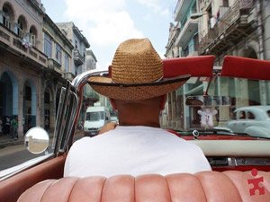 Persoon met strohoed rijdt in een vintage rode cabrio over een stadsstraat in Cuba