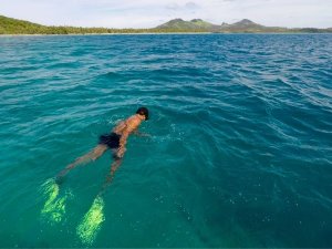 Snorkelaar met gele vinnen zwemt in helder blauw tropisch water met een groen eiland en bergen op de achtergrond aan de kust