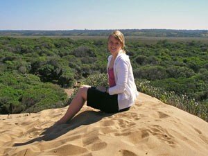A young woman sits on a sand dune overlooking a vast green forest.