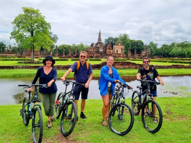 Vier mensen met fietsen staan op een grasveld voor een vijver. Op de achtergrond zijn oude tempelruïnes en stoepa’s te zien, omringd door bomen. De lucht is bewolkt.