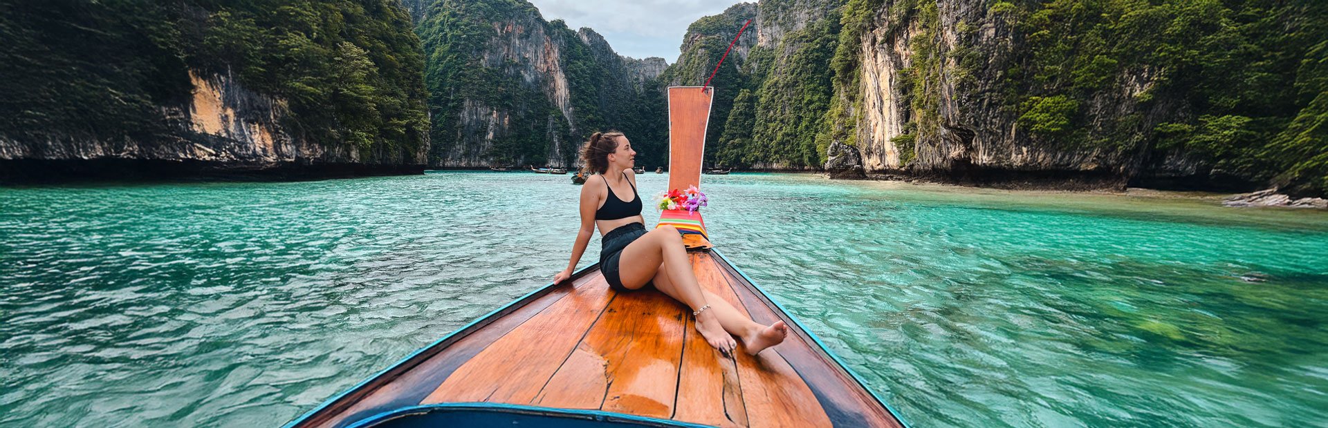 Vrouw ontspant op de punt van een traditionele longtailboot in helder turquoise water, omringd door groene kalkstenen kliffen bij Koh Phi Phi, Thailand.