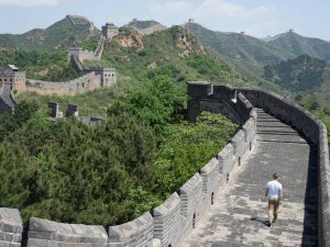 Een persoon springt in de lucht op een stenen pad langs de Chinese Muur, omringd door groene heuvels en wachttorens onder een blauwe lucht.