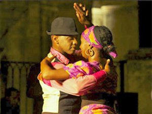 Man met zwarte fedora en vrouw met kleurrijke hoofddoek dansen dicht tegen elkaar aan in Cuba