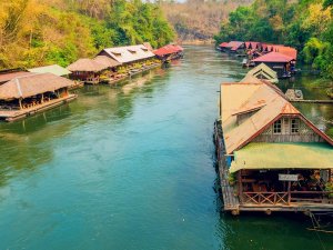 Houten huisjes met rieten of metalen daken drijven aan beide kanten van een brede rivier, omringd door groene bomen en heuvelachtige jungle.