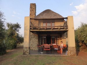 Vrouw zit op het schaduwrijke terras van een rustieke lodge met twee verdiepingen, een rieten dak en stenen schoorsteen in Zuid-Afrika