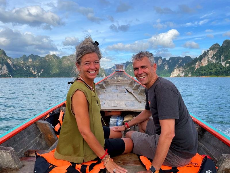 Twee reizigers zitten op een longtailboot op het water. Om hen heen rijzen steile kalksteenrotsen omhoog, begroeid met jungle. De lucht is blauw met een paar wolken, en op de houten boot liggen oranje reddingsvesten.
