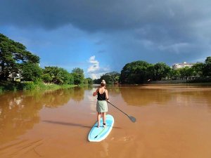 Reiziger staat op een blauw suppboard met een peddel in haar handen. De rivier is bruin van kleur, omringd door veel groen en een licht bewolkte lucht.