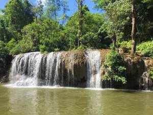 Een lage, brede waterval stroomt in een rivier, omringd door dichtbegroeid tropisch bos met groene bomen en struiken. Helder blauwe lucht te zien door het blader dek van de bomen.