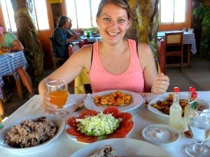 Blije jonge vrouw in een roze top zit aan een restauranttafel vol gerechten en een drankje en lacht naar de camera in Cuba
