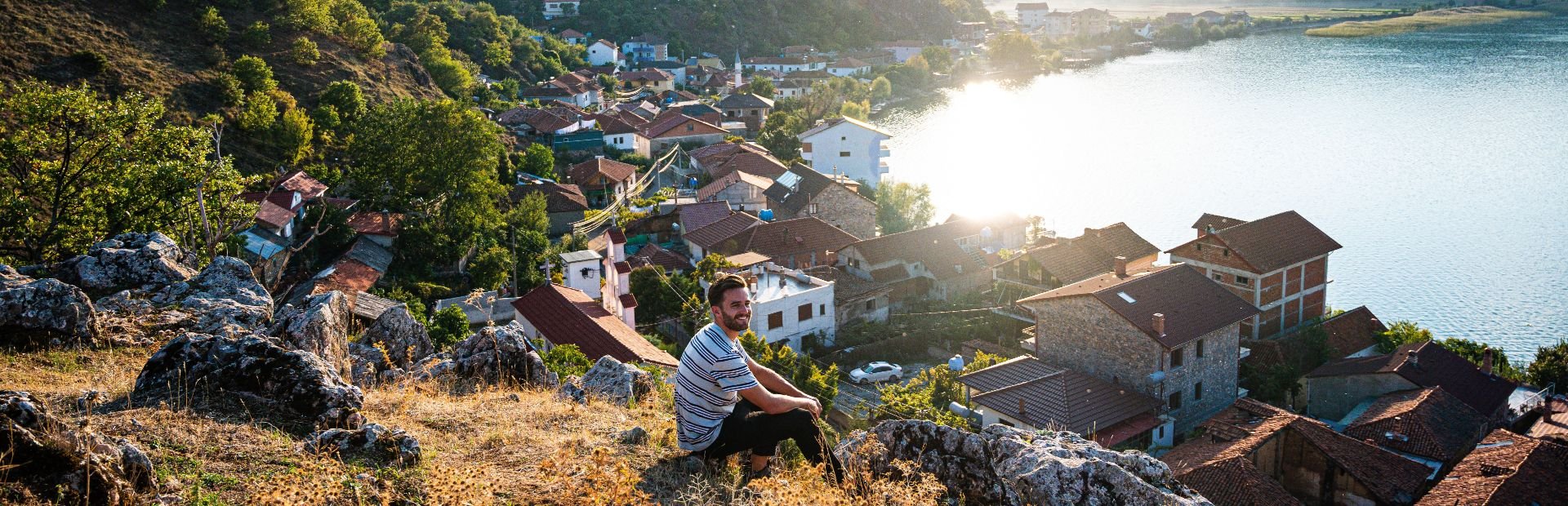 Man op een rotsachtige heuvel bij het Ohridmeer in Noord-Macedonië met uitzicht op een dorp en het zonovergoten meer