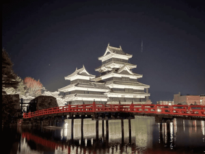 Verlicht Matsumoto-kasteel in Japan met rode brug ernaartoe, gereflecteerd in het water tijdens de nacht.