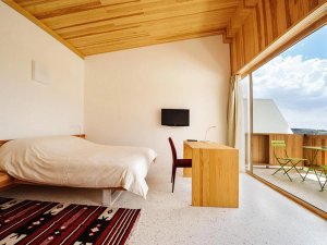 A modern minimalist bedroom with a sloped wooden ceiling, a bed, a desk, and a balcony with green chairs.