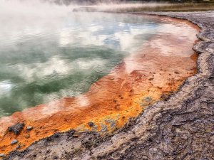 Stomende groene geothermische poel met oranje en rode mineraalafzettingen langs een rotsachtige oever in een vulkanisch natuurgebied