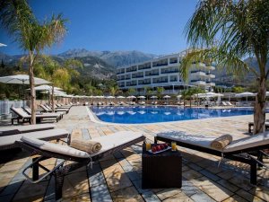 Outdoor resort swimming pool area with lounge chairs, palm trees, a white hotel building, and mountains under a clear sky.