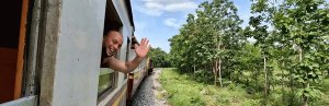 Reiziger zwaait lachend uit het raam van een trein die door het groene Thaise landschap rijdt