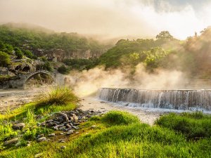 Mistig rivierlandschap in Albanië met een oude stenen boogbrug en een kleine waterval, omringd door groene heuvels in zonlicht