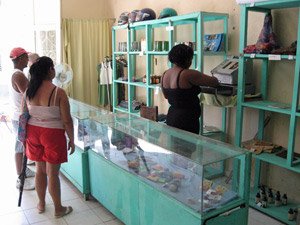 Vrouw achter de toonbank in een winkel bedient een weegschaal terwijl twee klanten wachten met schappen vol producten op de achtergrond in Cuba